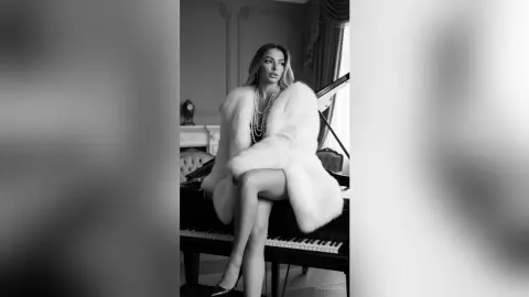 Elegant woman in a white faux fur coat sitting on a grand piano in a moody, sophisticated black and white setting.