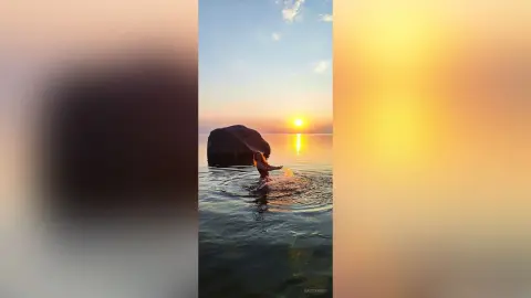 Redheaded woman wading in calm ocean water during a golden sunset with a large rock in the background.