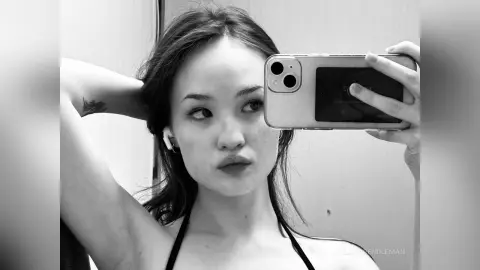 Young woman in black bikini taking a moody black and white mirror selfie in a bright bathroom.
