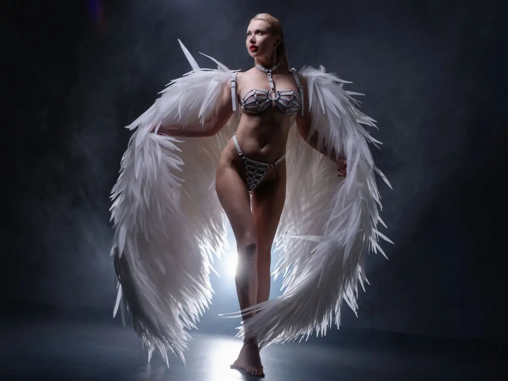 Blonde woman in white angel wings and bikini posing in a moody, smoky studio setting.