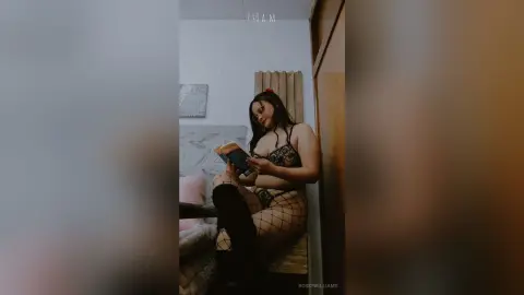 Young woman in black lace lingerie and fishnet tights reading a book in a cozy, moody bedroom setting.
