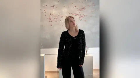 Blonde woman in a sparkly black cardigan and black trousers posing elegantly against a patterned wall.