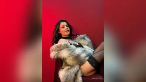 Glamorous woman in a black lace outfit and grey faux fur coat posing against a vibrant red background.