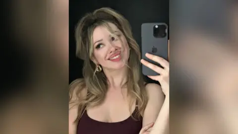 CassieArmas smiles for a mirror selfie, wearing a dark maroon tank top with her blonde hair styled in loose waves against a black background.
