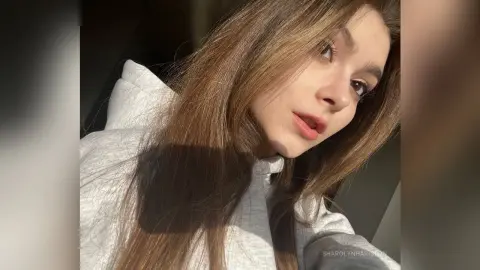 Young woman with long brown hair wearing a cozy white hoodie in soft natural sunlight.