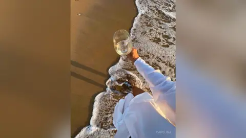 Person in a white linen outfit holding a glass of white wine by the ocean waves at sunset.