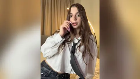 GertudeCova poses in a white button-down shirt and black tie, wearing black denim pants and long pink nails against a neutral curtain background.