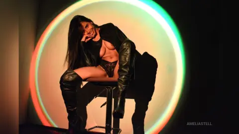 AmeliaStell poses sitting on a stool wearing black boots and black lace panties, framed by a large glowing ring light.