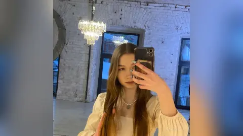 MagalyBoffa poses for a mirror selfie wearing a white long-sleeved top and a pearl necklace, with her long brown hair down in a bright room with a chandelier.