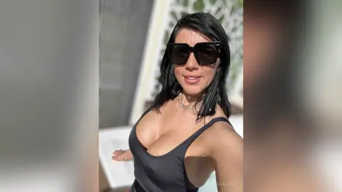 Smiling woman in a black tank top and sunglasses posing outdoors in bright sunlight.