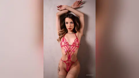 Glamorous woman in a pink lace bodysuit posing against a neutral wall with a confident mood.