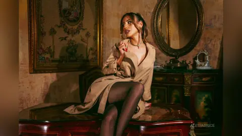 Elegant woman in a beige trench coat and black tights sitting in a vintage luxury room with gold decor.
