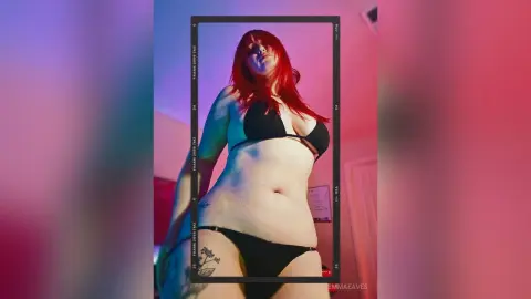 Redheaded woman in black bikini posing in moody red and blue lighting.