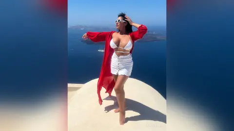 MissyJolie stands outdoors in a white bikini top and white skirt with a long red open robe, posing against a blue ocean backdrop.