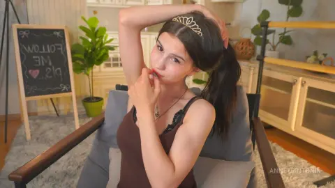 Young woman in a brown tank top wearing a tiara, sitting relaxed in a cozy living room setting.