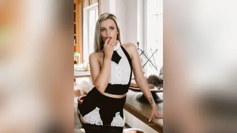 Gia poses in a white and black halter top and matching skirt, resting one hand near her mouth while looking at the camera.