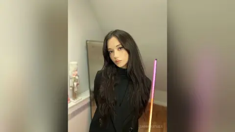 SabrinaSteels poses with long, wavy dark hair, wearing a black turtleneck in a room with a mirror and soft lighting.