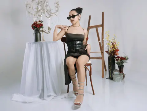 BrianaKnox — Stylish woman in a black satin dress and sunglasses sitting by a floral table in a bright studio setting.