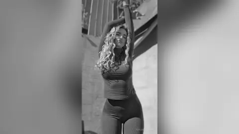 TiffanyAddams poses outdoors in black and white, wearing a tight long-sleeve top and leggings with her arms raised above her head.