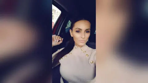 AishaApril poses in a tan turtleneck top with gold button details, looking directly at the camera while sitting in a car.