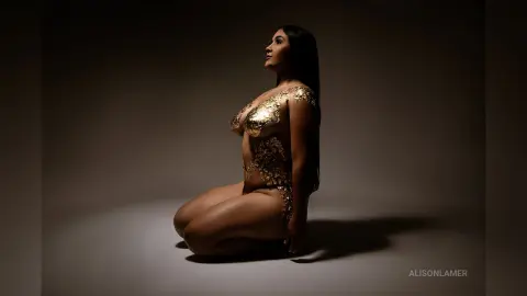 Elegant woman in a shimmering gold sequin bodysuit posing in a moody, dark studio setting.