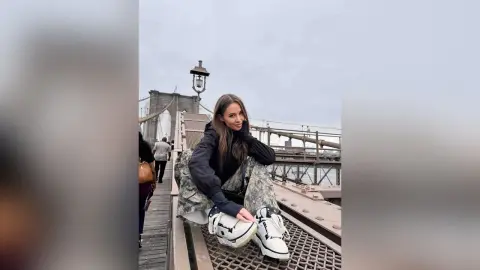 EvelynMills sits on a metal walkway of a bridge wearing a black hoodie, camouflage shorts, and white sneakers.