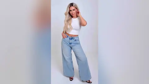 Blonde woman in a white crop top and light wash wide-leg jeans posing against a white studio background.