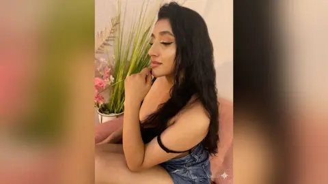 Young woman in a black top and denim shorts posing thoughtfully near indoor plants in a bright room.