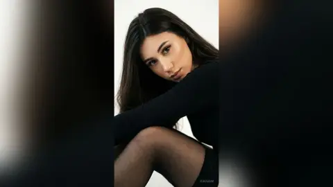 Young woman with long dark hair wearing a black long-sleeve top in a minimalist studio setting with a moody vibe.
