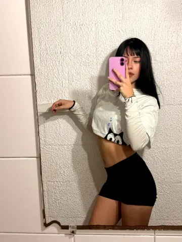 Young woman in a white long-sleeve crop top and black mini skirt taking a mirror selfie in a modern bathroom.