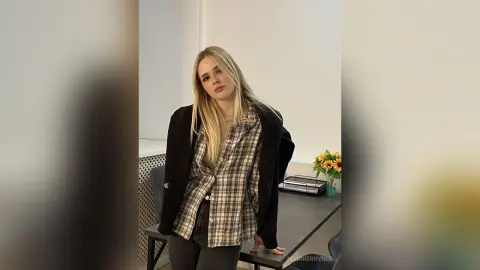 Blonde woman in a plaid shirt and black blazer posing confidently in a bright modern office setting.