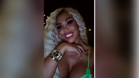 Glamorous blonde woman in a green top wearing a gold watch, smiling in low light evening setting.