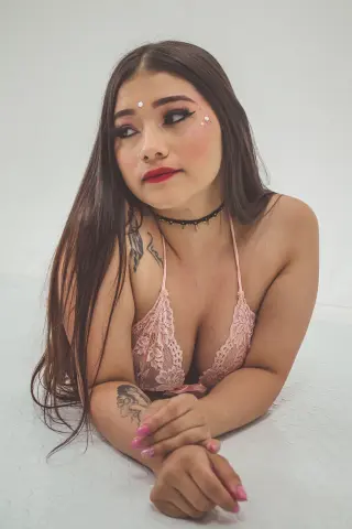 Young woman with long dark hair and tattoos wearing a pink lace bikini top in a bright studio setting.