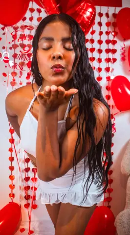 Cheerful woman in a white summer outfit blowing a kiss amidst red heart decorations and balloons.