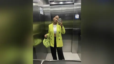 MissyJohansson poses in a mirror selfie inside an elevator, wearing a bright lime green blazer, a white crop top, and black pants while holding a green bag.