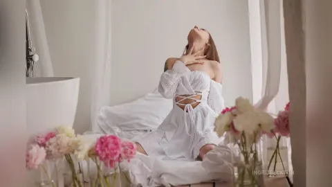IngridBeckton poses on a white bed, wearing a white lace-up top, with her head tilted back and hands touching her neck.