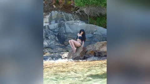 Young woman in black bikini sitting on rocks by the ocean waves in a serene coastal setting.