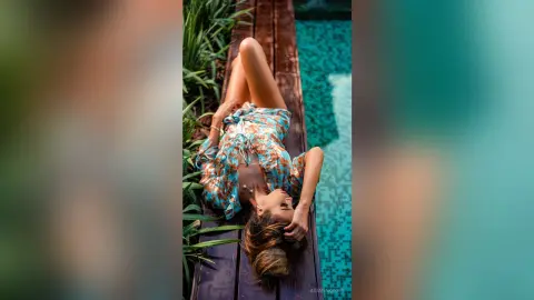Relaxed woman in a floral summer dress lounging on a wooden deck by a turquoise swimming pool.