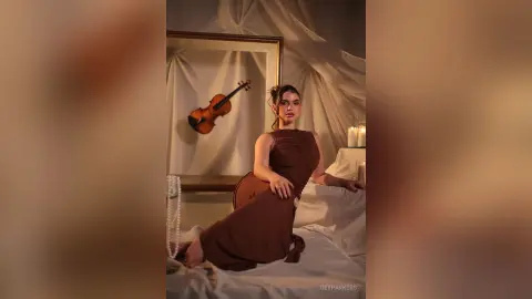 Elegant woman in a brown cocktail dress sitting on a bed in a moody, dimly lit room with a violin decor.