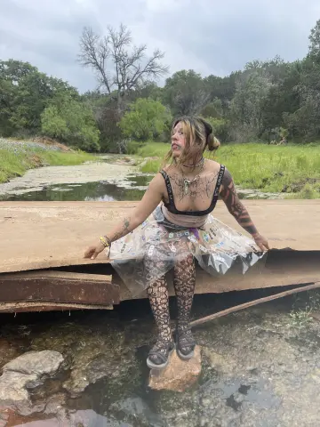 Bohemian woman in a sheer patterned skirt and tank top standing by a tranquil forest creek on a cloudy day.