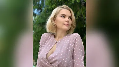 Blonde woman in a pink polka dot blouse smiling peacefully in a lush green garden setting.