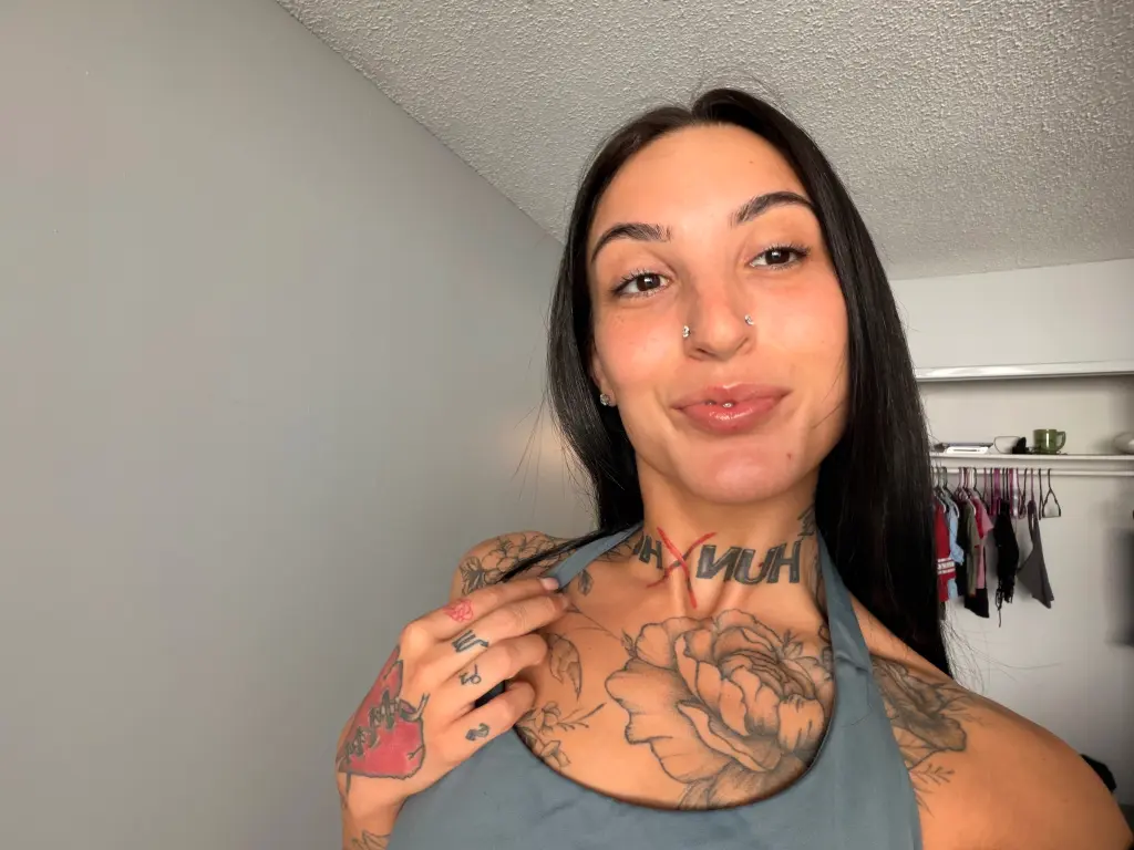 DylanAshley is a smiling woman with long dark hair and tattoos on her chest and shoulder, wearing a grey tank top.