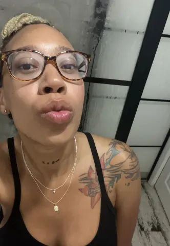 Woman with glasses and tattoos wearing a black tank top in a modern indoor setting.