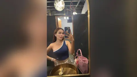 Young woman in a navy blue tank top taking a mirror selfie in a modern bathroom with warm lighting.
