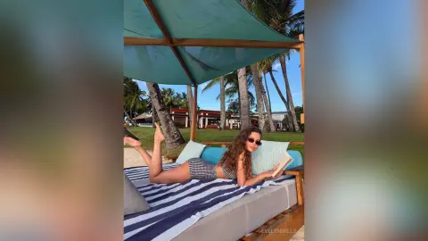 Relaxed woman in a black bikini reading a book on a lounge bed under a tropical beach canopy.