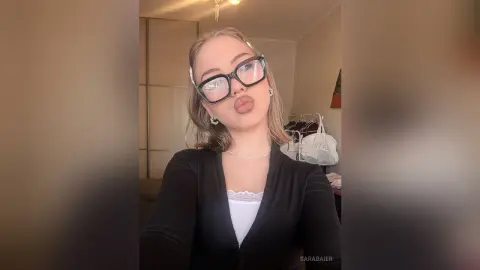 SaraBaier wearing black glasses and a black top while making a pouty face at the camera.