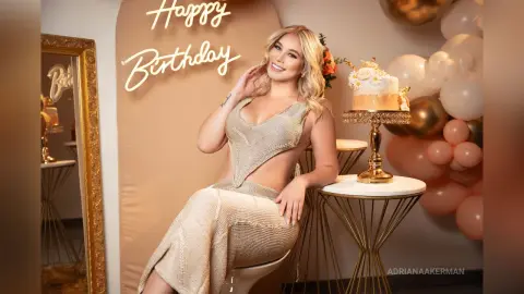 Smiling woman in a sparkly gold dress celebrating a birthday with balloons and a cake in a festive setting.