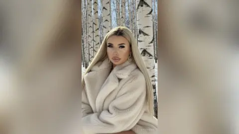Blonde woman in a luxurious white faux fur coat posing in a snowy birch forest setting.