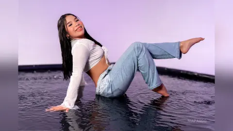 Smiling young woman in a white long-sleeve crop top and light blue jeans sitting playfully on a dark water surface.