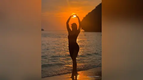 AnaMathers stands in silhouette on a beach at sunset, holding her hands up to form a heart shape against the sun.